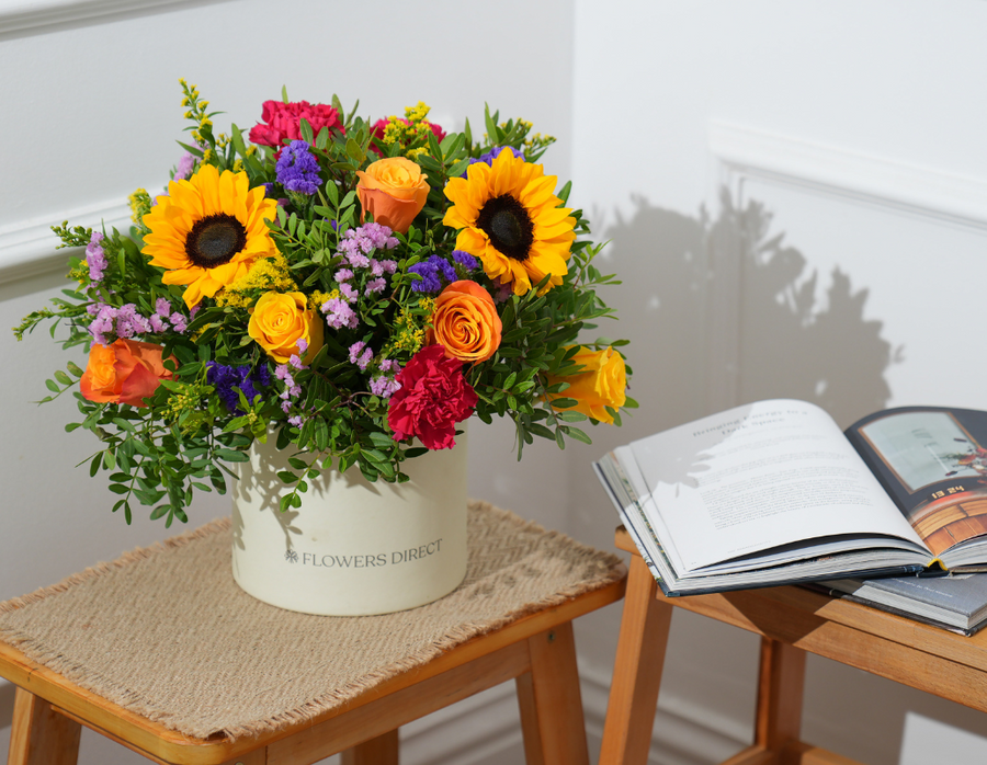 Vibrant summer flower hatbox with sunflowers, orange roses, pink carnations and purple statice on side table with open book
